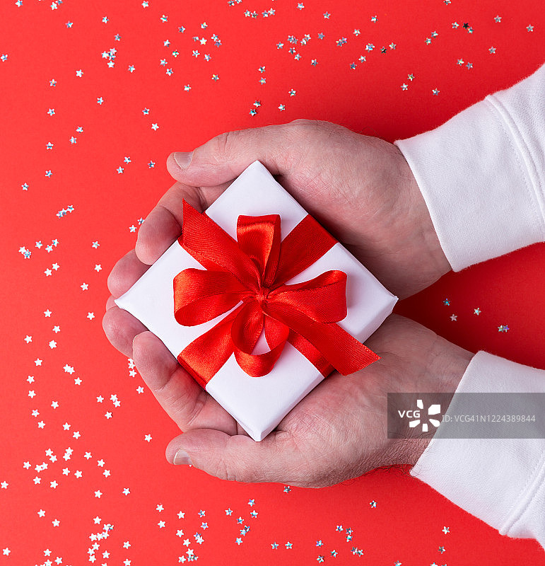 礼盒包装在白色的纸与一个红色的蝴蝶结在一个红色的背景拷贝空间。男人手中的情人节礼物。圣诞礼物，给女人的礼物。生日礼物。图片素材