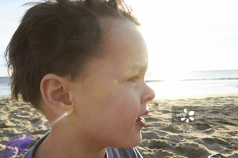 小男孩棕色短卷发棕色眼睛侧面他的脸近海滩和阳光在他的背景他正在眯着眼睛图片素材