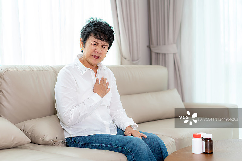亚洲老年妇女在他的胸部不适心脏病发作与痛苦的胸痛在客厅在家里。图片素材