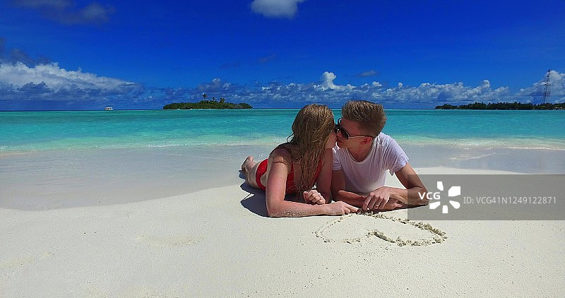 在一个阳光明媚的日子里，一对夫妇在美丽的白色沙滩上画了一颗心，蓝色的天空和云的背景图片素材