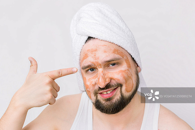 一个英俊的微笑轻松的年轻人的肖像与一个面部泥粘土面具，指向他的脸。面部和身体皮肤护理治疗。孤立在白色图片素材