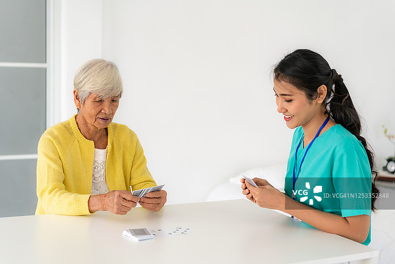 亚洲年轻女性护士或家庭健康助理与老夫妇打牌在客厅的桌子在家里的家庭保健概念。图片素材