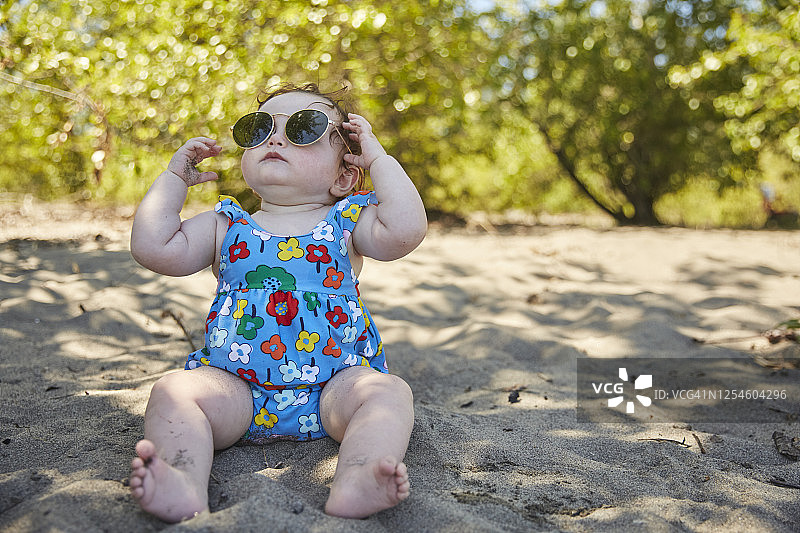 一个可爱的婴儿坐在沙滩上，脸上戴着墨镜。图片素材
