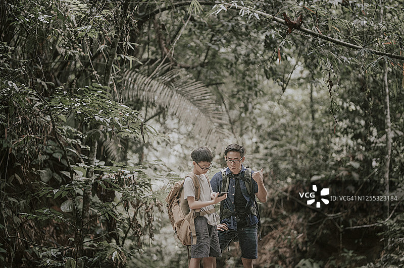 一位亚洲华人父亲带着儿子在森林里徒步旅行和探索，他正在看地图上的手机应用程序来导航图片素材