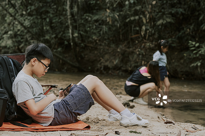 一个亚洲的中国男孩独自坐在河边玩手机游戏，而他的妹妹和妈妈周末在森林里的河里玩图片素材