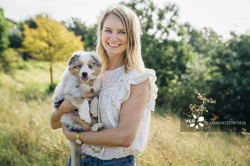 一幅微笑的金发女子在公园外抱着澳大利亚牧羊犬小狗的肖像图片素材