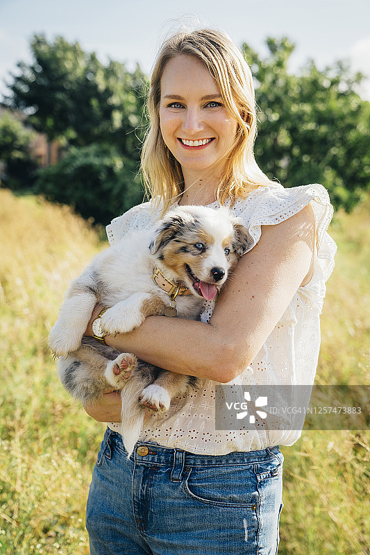 一幅微笑的金发女子在公园外抱着澳大利亚牧羊犬小狗的肖像图片素材