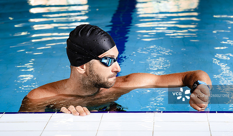 一位专业游泳运动员站在游泳池里，一边观察手上的防水手表，一边评估自己游泳的时间。图片素材