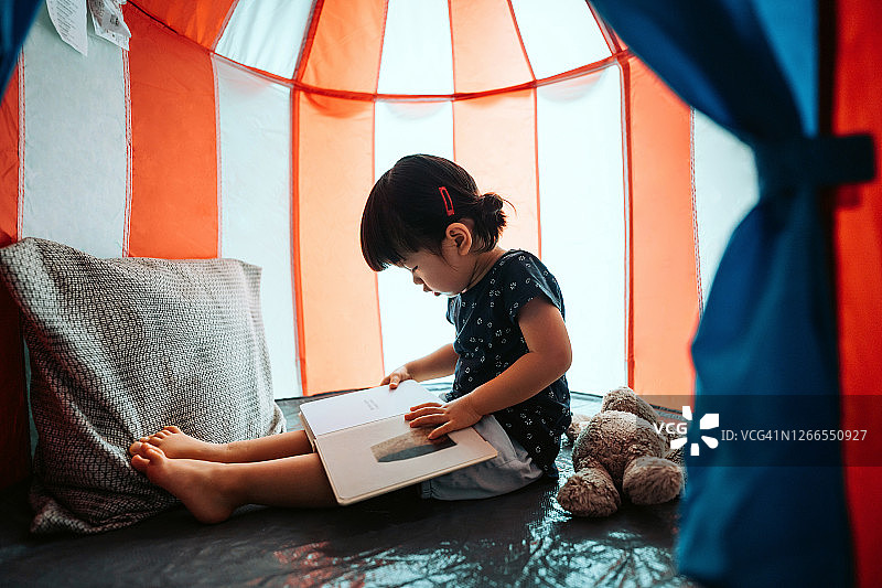 可爱的小亚洲女孩享受自己和花闲暇时间在家里。她正在家里的帐篷里和她的泰迪熊一起读书图片素材
