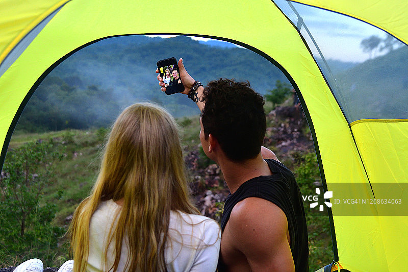 一对年轻情侣坐在帐篷里露营，男友用智能手机自拍图片素材