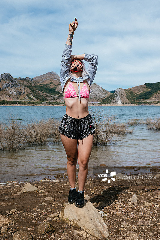 年轻女子在西班牙的一个湖边，穿着比基尼和短裤。双臂向上，以放松的姿势站立图片素材