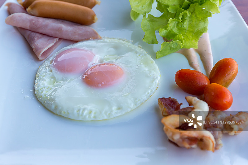 美式早餐:香肠，鸡蛋，火腿，培根和蔬菜放在白色的盘子里。传统早餐有煎蛋和香肠。木桌上放着早餐的煎蛋、香肠和火腿。图片素材