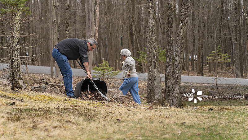 一个成熟的男人在帮助他的母亲，一个年长的女人，清理乡间房子后院的老树叶。图片素材