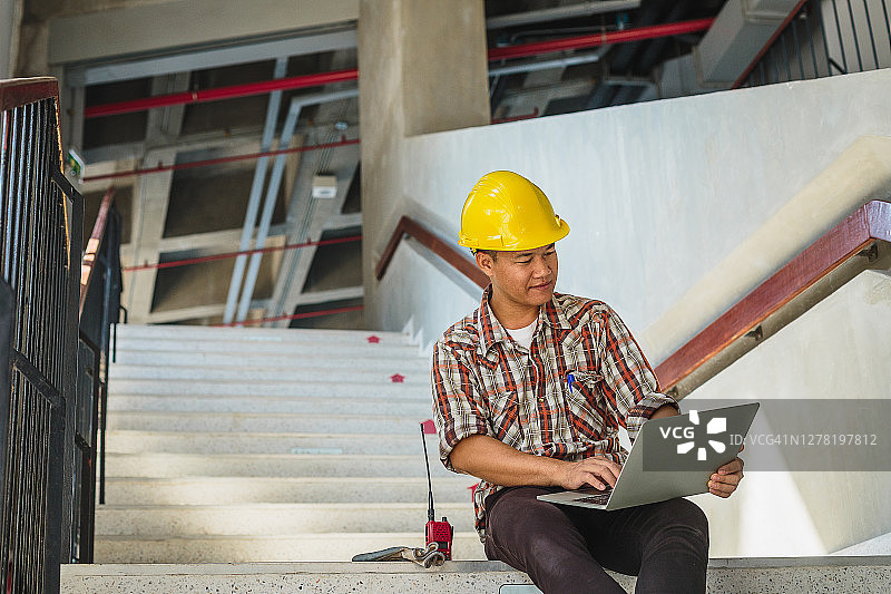 亚洲工程师戴安全帽在工地楼梯上使用笔记本电脑，工程师在工地检查建筑工程图片素材
