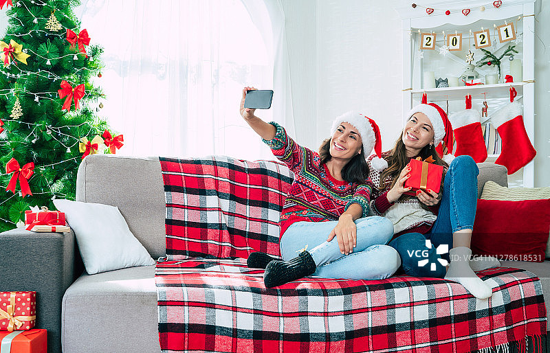 新年快乐，圣诞快乐。两个戴着圣诞帽的年轻美丽的姐妹或朋友正在沙发上用智能手机与圣诞礼盒自拍。图片素材