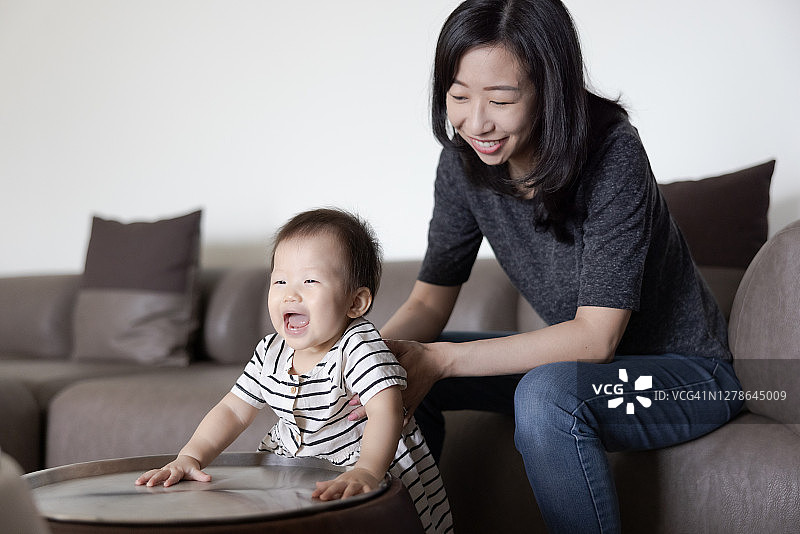 可爱的亚洲宝宝和她的妈妈在客厅的沙发旁边快乐地玩耍图片素材