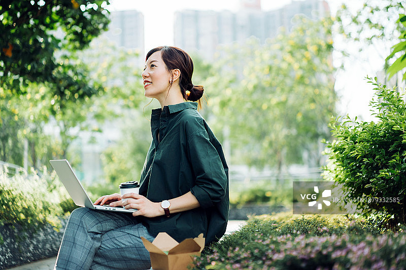 一位自信的年轻亚洲女商人坐在城市公园的长椅上，一边喝着咖啡，一边拿着健康沙拉午餐盒，在午休时间用笔记本电脑工作图片素材