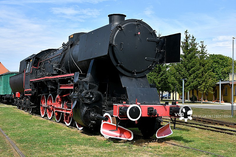 近距离观看一列老式火车，黑色金属车身，红白相间的车轮停在旁边的铁轨上，铁轨周围环绕着树木，背景是波兰的火车站图片素材