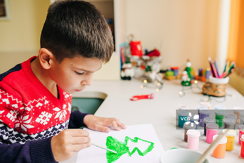 在他的育儿室的桌子上，一个小男孩在一张白色的床单上画着一棵圣诞树图片素材