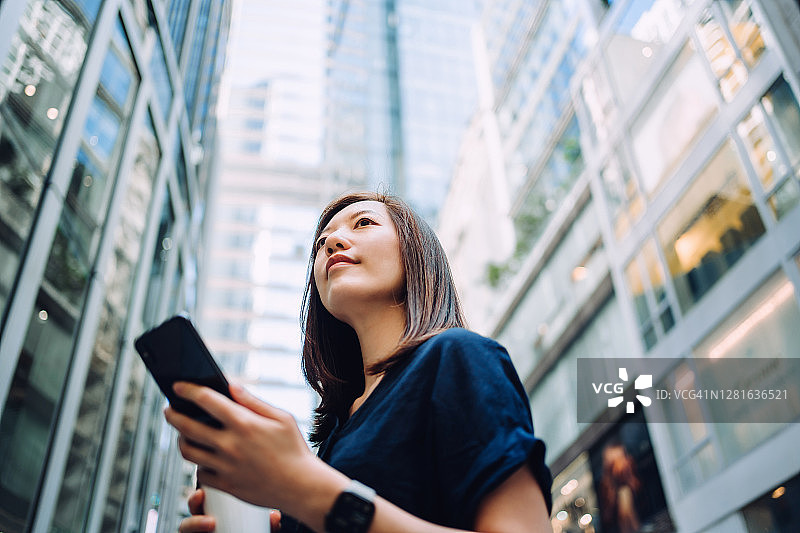 这是一幅自信的年轻亚洲女商人的低角度肖像，她一边喝着咖啡，一边用智能手机站在这座城市金融区的高楼大厦前图片素材