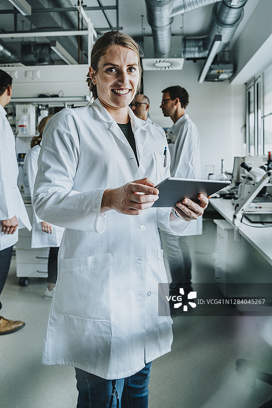 在实验室里，一名微笑着使用平板电脑的女性，站在同事身后图片素材