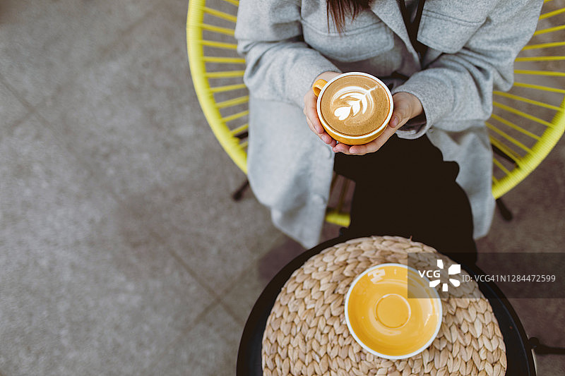 陌生女人的手拿着黄色的咖啡杯。女人的手在温暖的秋天衣服拿着卡布奇诺而坐在户外咖啡馆的黄色椅子的模型图像。平躺，俯视图。图片素材