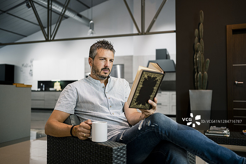 放松的男性自由职业者阅读书籍，而坐在办公室的椅子上与咖啡杯图片素材