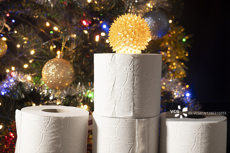 厕纸卷上有一个尖球，可能代表新冠病毒。背景上装饰着彩灯的圣诞树。图片素材