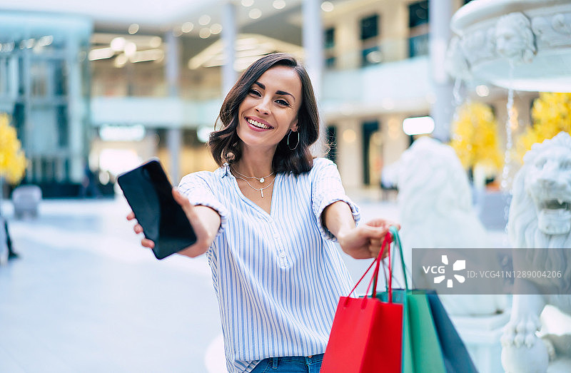 在黑色星期五，年轻漂亮时髦的女人拿着购物袋在商场里玩智能手机图片素材
