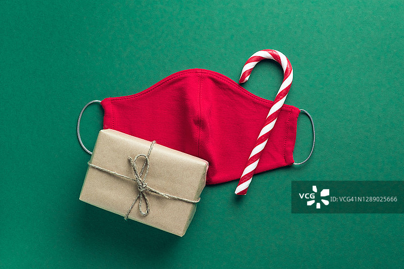 红色医用防护口罩，拐杖状条纹糖果，绿色圣诞背景的礼物。预防和预防COVID - 19大流行的传播。一个快乐健康的圣诞节的概念。图片素材