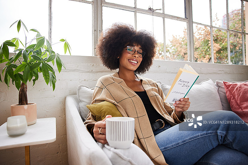 微笑的年轻女子放松在她的沙发与一本书和一杯咖啡图片素材