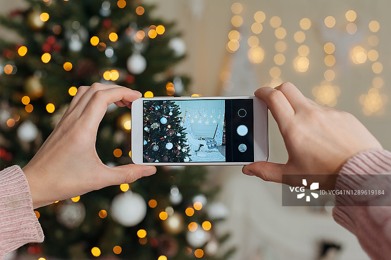 女孩拍摄智能手机模型的照片视频。在新年和圣诞节的背景下，一个舒适的室内装饰着一棵圣诞树。图片素材