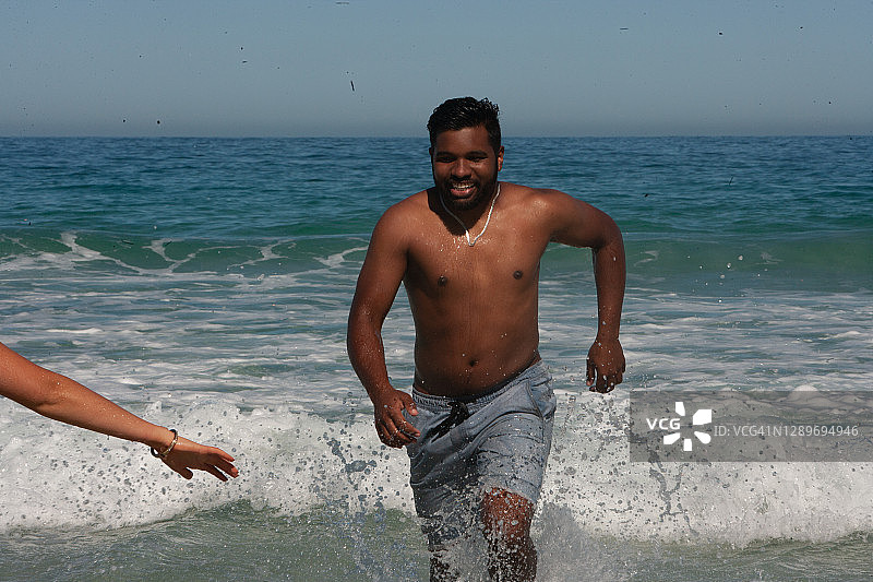 一个英俊快乐的年轻人跑出海滩的海浪迎接他的伴侣伸出的手在等待图片素材