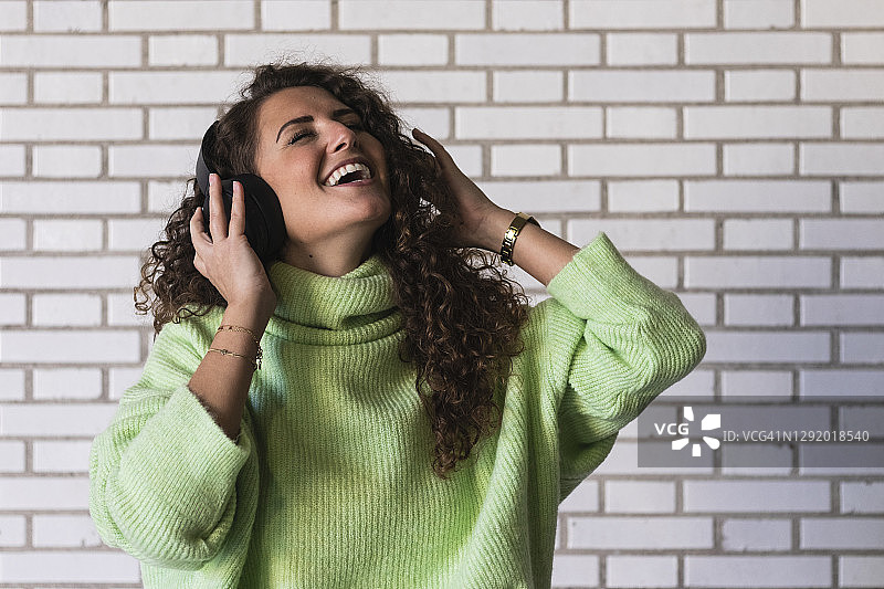 一名年轻女子靠在砖墙上，戴着耳机听音乐图片素材