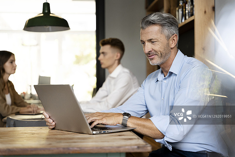 一个成熟的商人在办公室和同事坐在一起用笔记本电脑工作图片素材