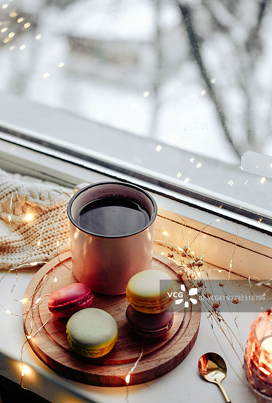 热饮咖啡在粉红色的杯子，童话灯，美味多彩的饼干马卡龙在木制托盘，窗户和下雪的天气，冬天舒适的圣诞图片。图片素材