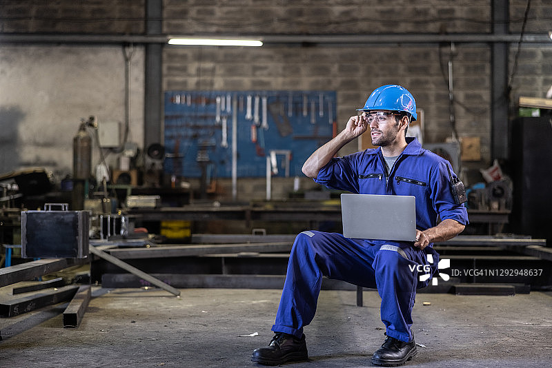 白人工人穿着蓝色工作服和头盔，在工厂内部使用笔记本电脑图片素材