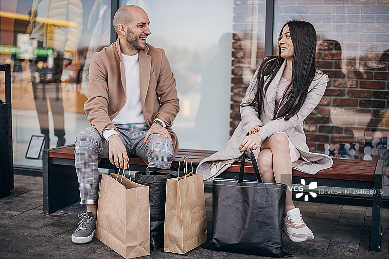 两个年轻人购物后拿着购物袋坐在长凳上图片素材