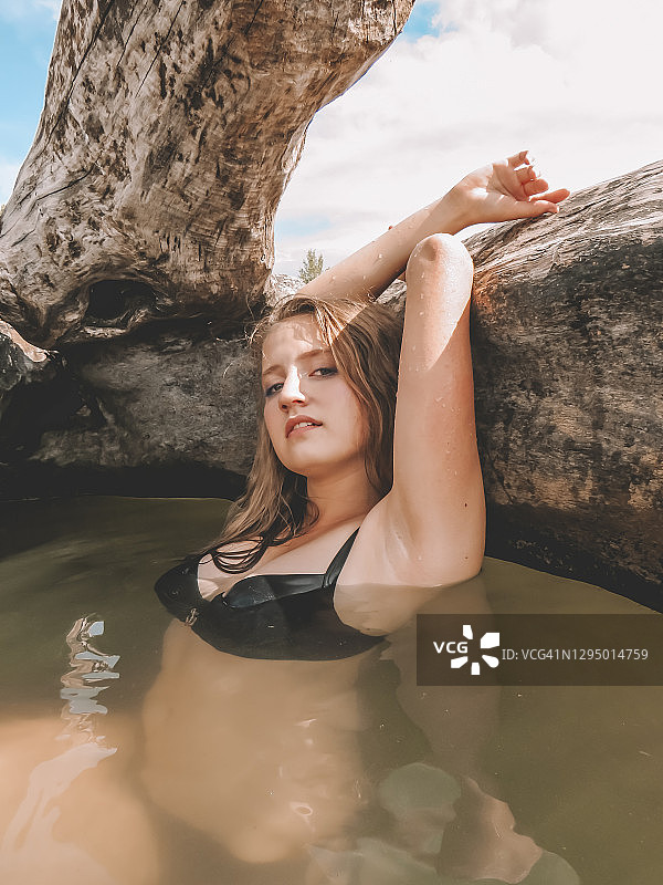 红头发的年轻女子与松散的头发在一个炎热的夏天，在一个巨大的干树的树干在海湾的水一个美丽的身体图片素材