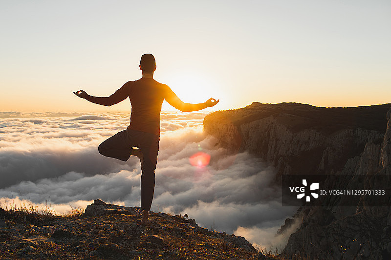 在日落时分，一个人在天空之上的山上独自练习哈他瑜伽。轮廓从后面图片素材