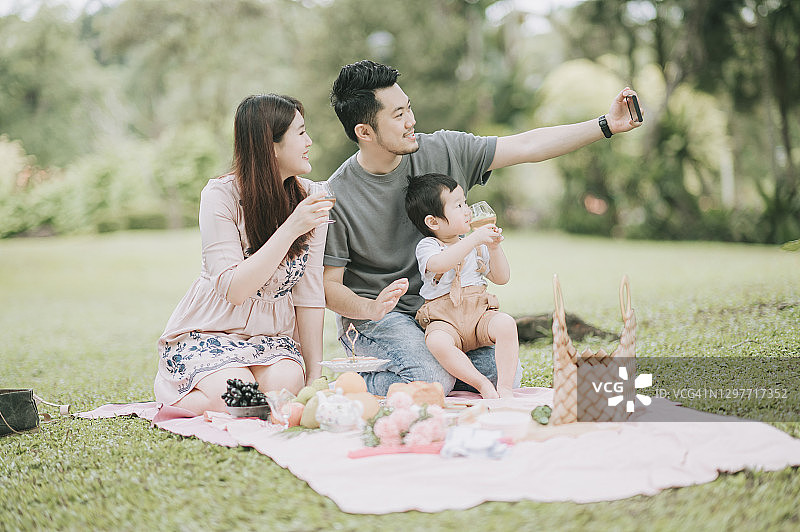亚洲华人年轻家庭使用智能手机自拍享受户外野餐时间在周末图片素材