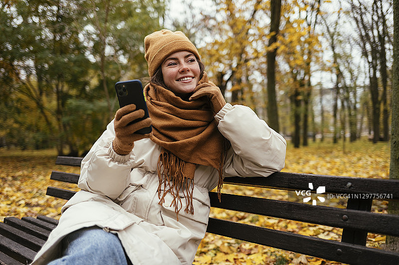 微笑的女人在秋天的公园里拿着手机做白日梦图片素材