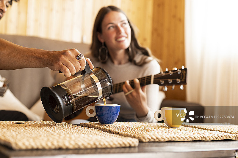 男朋友往杯子里倒咖啡，而女朋友坐在家里弹吉他图片素材