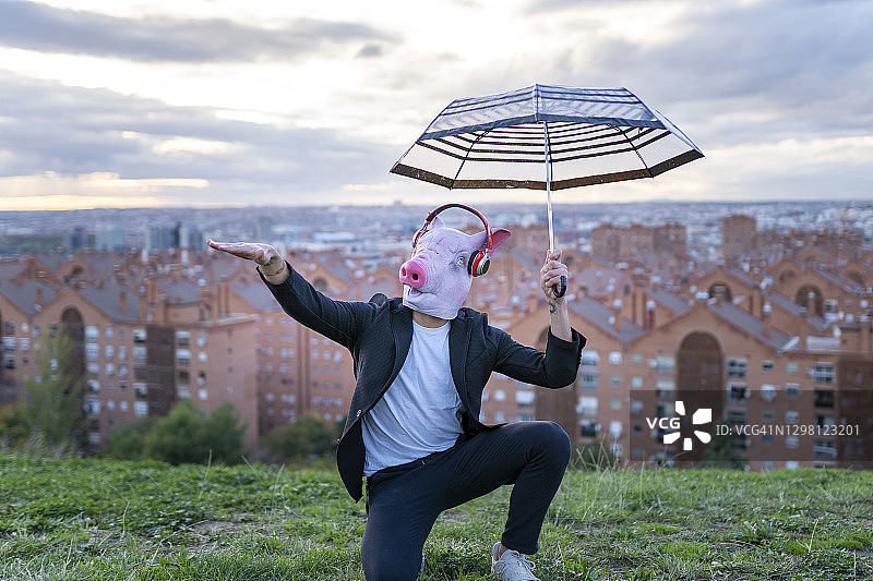 男专业人士戴着猪口罩，撑着伞，在日落时跪在天空下打手势图片素材