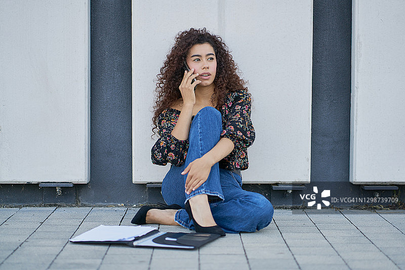在户外打电话的女人。一个年轻女子坐在户外用手机聊天和阅读一些文件的肖像。图片素材