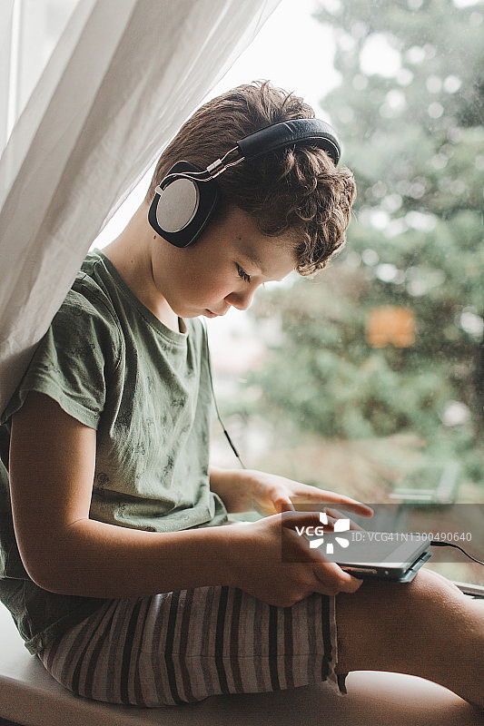 小男孩坐在窗台上，带着平板电脑和耳机听音乐玩游戏图片素材