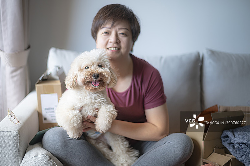 亚洲华人短发中年妇女与她的宠物玩具贵宾犬在沙发上看着照相机开心的微笑图片素材