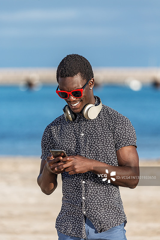 欢快时髦的黑人戴着耳机在海滩上玩智能手机图片素材