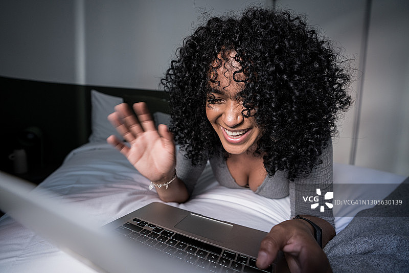 一名年轻女子躺在家里的床上用笔记本电脑进行视频通话时挥手图片素材