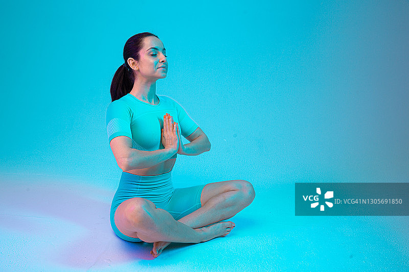 年轻运动有吸引力的女人练习瑜伽坐在莲花体位，穿着运动服，裤子和上衣，白色瑜伽隔离在霓虹灯背景梯度工作室。图片素材